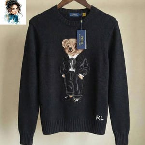 Może przedstawiać: Czarny sweter z misiem w garniturze wyszytym z przodu. Sweter ma prążkowany okrągły dekolt i mankiety. Widoczna jest metka z marką. Litery "RL" są wyszyte w prawym dolnym rogu. Sweter wisi na drewnianym wieszaku.