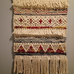 May include: A woven wall hanging with a cream-colored background and geometric patterns in red, black, and yellow. The wall hanging has a fringe at the top and bottom. The design includes diamond and triangle shapes.