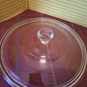 May include: A clear glass lid with a small, rounded handle in the center. The lid has a circular shape with a raised rim and a textured pattern on the surface. The background is a mix of purple and yellow.