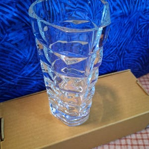 May include: A clear, faceted glass vase with a geometric design. The vase is cylindrical with a wide opening and sits on a brown cardboard box. The background is a blue fabric with a pattern.