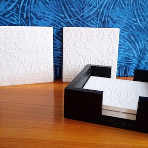 May include: White square coasters with an embossed design of a reindeer and trees. The coasters are displayed with a black wooden holder, and two additional coasters are standing upright. The background is a blue fabric.
