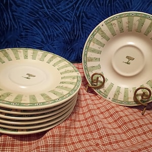 May include: A set of white ceramic plates with a green and white striped border design. Each plate features a small, centered illustration. One plate is displayed on a decorative metal stand. The plates are stacked on a red and white checkered surface.