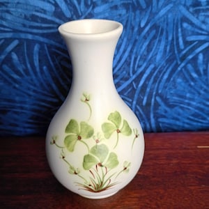 May include: A small, white ceramic vase with a flared top and rounded body. The vase is decorated with green and brown painted shamrocks. The vase is approximately 6 inches tall.