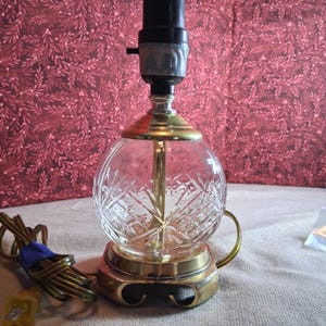 May include: A table lamp base featuring a clear, spherical glass body with a cut-glass crosshatch pattern. The lamp has a brass-toned metal base and neck, a black socket, and a brown electrical cord with a standard two-prong plug attached.