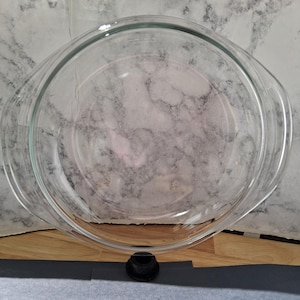 May include: A clear, round glass baking dish with integrated side handles. The dish features a smooth, transparent surface and a wide, open top design, suitable for oven use. The glass is entirely colorless and reflects light across its curved rim.