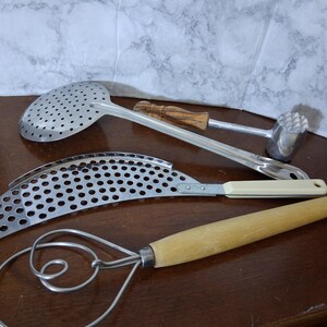 May include: A collection of three kitchen utensils: a stainless steel skimmer with a wooden handle, a curved metal strainer with a cream-colored handle, and a metal meat tenderizer featuring a textured head and a wooden handle.