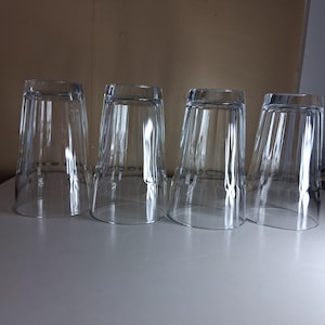 May include: A set of four clear glass tumblers positioned upside down in a row. Each tumbler features a tapered cylindrical shape with vertical ribbed detailing along the sides and a smooth, circular rim at the base for a simple, functional design.