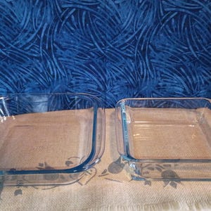 May include: Two clear, rectangular glass baking dishes. The dishes have rounded corners and a slight blue tint. The larger dish is on the left, and the smaller dish is on the right. The background is a blue fabric with a white pattern.