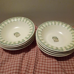 May include: A set of cream-colored soup bowls with a green and white striped border and a small cabin illustration in the center. The bowls are stacked in two piles on a red and white checkered tablecloth.