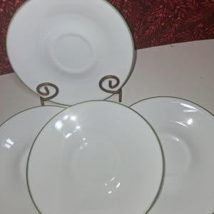 May include: A set of four white ceramic plates with a light green trim. One plate is displayed on a decorative metal stand. The plates have a simple, elegant design, suitable for everyday use or special occasions.