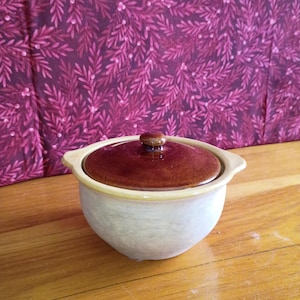 May include: A small, ceramic casserole dish with a lid. The dish is off-white with a tan rim and a dark brown lid. The lid has a small, round knob. The background is a patterned fabric in shades of pink and purple.