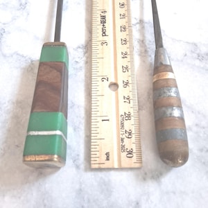 May include: Two metal tools with decorative handles placed next to a wooden ruler. The left handle features green resin and dark wood segments with a thin silver band. The right handle has alternating wood and silver-toned metal rings.