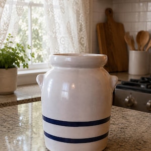 May include: A white ceramic crock featuring a wide mouth, a rounded body, and two small side handles. The exterior is decorated with two horizontal navy blue stripes encircling the lower portion of the vessel, providing a simple contrast.