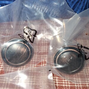May include: Two bronze-colored cabinet knobs in clear plastic packaging. The knobs are round with a slightly raised center and a dark, aged finish. The packaging is sealed and ready for use.