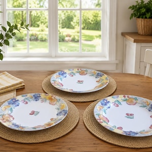May include: Three white ceramic dinner plates featuring a colorful watercolor-style fruit pattern around the rim and a small cherry motif in the center. Each plate rests on a round, woven tan placemat on a wooden table surface.