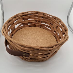 May include: A round, open-weave brown wicker basket featuring a flat, circular cork base insert. The basket is constructed with thick, flexible woven reeds and includes a single dark brown leather handle attached to the side with a metal rivet.