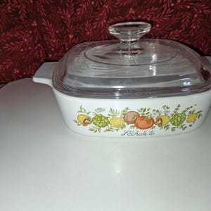 May include: A white rectangular glass casserole dish with a clear glass lid and integrated side handles. The exterior features a colorful botanical print of vegetables and herbs with the text L'Echalote printed in black script on the front side.