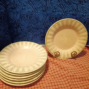 May include: A stack of six off-white ceramic plates with a green striped border. One plate is displayed on a decorative metal stand. The plates are on a red and white checkered tablecloth, with a blue patterned background.