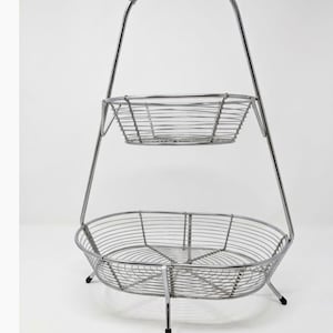 May include: A two-tiered wire basket stand constructed from polished chrome metal. The unit features a smaller upper basket and a larger lower basket, both with a circular wire grid design, supported by a central frame with a coiled handle.