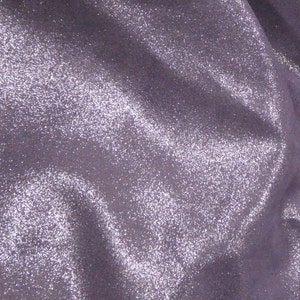 Puede incluir: Primer plano de una tela brillante de color lavanda. El material está cubierto de diminutas lentejuelas brillantes, creando una superficie texturizada y reflectante. La tela está drapeada, con pliegues suaves y sombras.