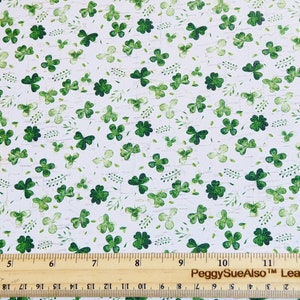 May include: A white sheet of material featuring a repeating pattern of green shamrocks and small sprigs. A wooden ruler is positioned at the bottom, showing measurements in inches from 4 to 12 and centimeters from 10 to 21.