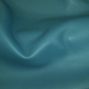 Puede incluir: Primer plano de cuero de color verde azulado con una superficie texturizada. El cuero presenta sutiles variaciones tonales, con áreas más oscuras que crean sombras y reflejos. El material es suave y flexible, con pliegues y arrugas visibles.