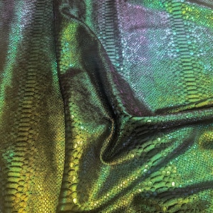 May include: Close-up of iridescent fabric with a snakeskin pattern. The fabric transitions from dark green to yellow and purple, with a shimmering, textured surface. The material is folded, creating shadows and highlights.
