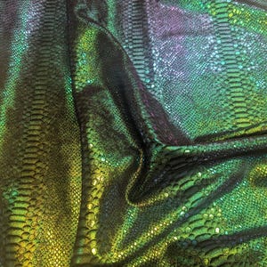 Puede incluir: Primer plano de una tela iridiscente con un patrón texturizado en forma de escamas. La tela transita del verde oscuro al amarillo y al morado, creando un efecto brillante. El material está doblado, mostrando las variaciones de textura y color.