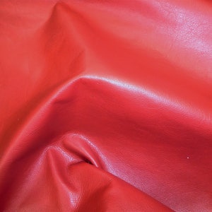 May include: A close-up of a vibrant red leather material. The leather has a smooth, slightly glossy finish with subtle variations in tone, creating a rich, textured appearance. The folds and creases in the material add depth and dimension to the image.