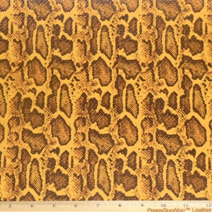 May include: Close-up of a textured fabric with a snakeskin pattern. The design features a repeating pattern of brown and tan scales. The fabric is marked with a ruler in inches.