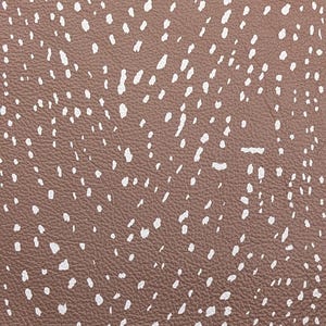 May include: Close-up of a brown leather material with a pattern of irregular white spots. The texture appears to be slightly pebbled, with the white spots varying in size and shape, creating a speckled effect.