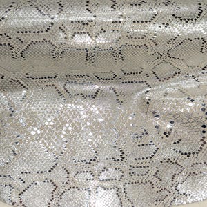 May include: Close-up of a silver snakeskin pattern. The fabric has a textured surface with a repeating geometric design. The pattern is a mix of silver and black, creating a shimmering effect. The material appears to be flexible and draped.