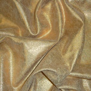 May include: Close-up of a shimmering gold fabric with a textured surface. The material is draped, creating soft folds and shadows. The fabric has a metallic sheen, reflecting light and adding a luxurious feel. Ideal for apparel, home decor, or craft projects.