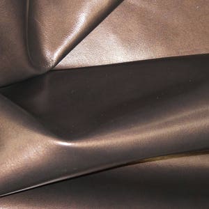 May include: Close-up of a dark brown leather material with a glossy finish. The leather is folded and creased, creating shadows and highlights that emphasize its texture and depth. The overall appearance is smooth and luxurious.