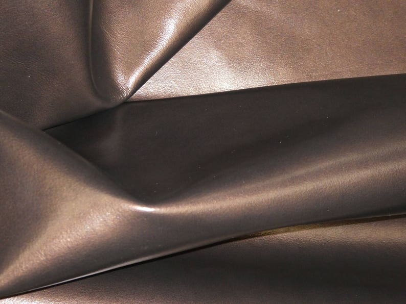 May include: Close-up of a dark brown leather material with a glossy finish. The leather is folded and creased, creating shadows and highlights that emphasize its texture and depth. The overall appearance is smooth and luxurious.