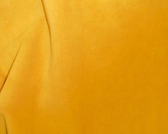 May include: Close-up of a textured, golden-yellow fabric with subtle folds and shadows. The material has a soft, velvety appearance, suggesting a luxurious textile. The color is consistent throughout, with variations in tone due to the light.