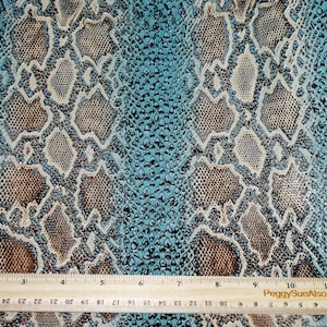 May include: Close-up of a turquoise blue fabric with a snakeskin pattern. The fabric has a textured, embossed look. A wooden ruler is placed at the bottom of the image.