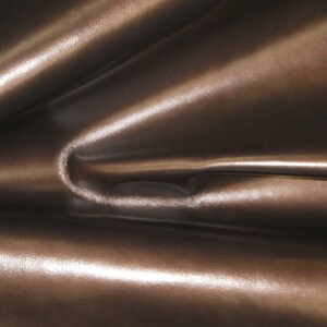May include: Close-up of a shiny, brown fabric with a metallic sheen. The material is folded, creating soft curves and highlights. The texture appears smooth and luxurious, suggesting a high-quality textile.