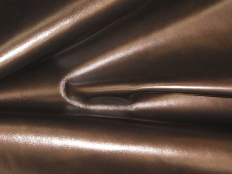 May include: Close-up of a shiny, brown fabric with a metallic sheen. The material is folded, creating soft curves and highlights. The texture appears smooth and luxurious, suggesting a high-quality textile.