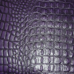 Puede incluir: Primer plano de un material de cuero sintético morado texturizado. La superficie tiene un patrón repetitivo de formas rectangulares y ovaladas, que se asemeja a la piel de cocodrilo o caimán. El color es un morado intenso y rico con sombras más oscuras en las grietas.