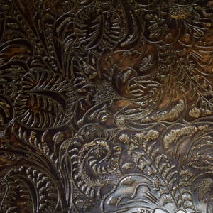 May include: Close-up of a brown leather surface with an embossed floral pattern. The pattern is intricate and detailed, with a metallic sheen.