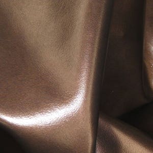 May include: Close-up of brown leather with a textured surface. The leather has a rich, dark brown color with subtle variations in tone. The folds and creases create shadows and highlights, adding depth and dimension to the material.