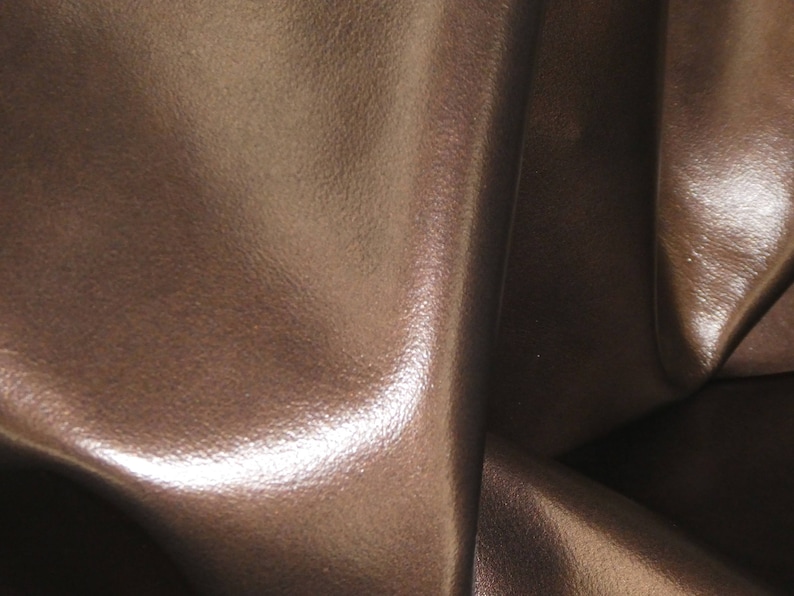 May include: Close-up of brown leather with a textured surface. The leather has a glossy sheen and appears to be folded, creating shadows and highlights. The color is a rich, warm brown.