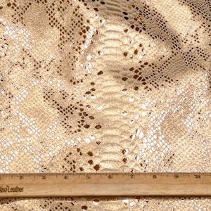 May include: Gold metallic fabric with a snakeskin pattern. The fabric is textured and has a subtle shimmer. A wooden ruler is included for scale.