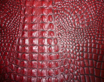 May include: Close-up of a textured, red leather surface. The pattern resembles crocodile or alligator skin, with a repeating scale-like design. The colour is a deep, rich red with subtle variations in tone, creating a realistic appearance. The texture appears slightly glossy.