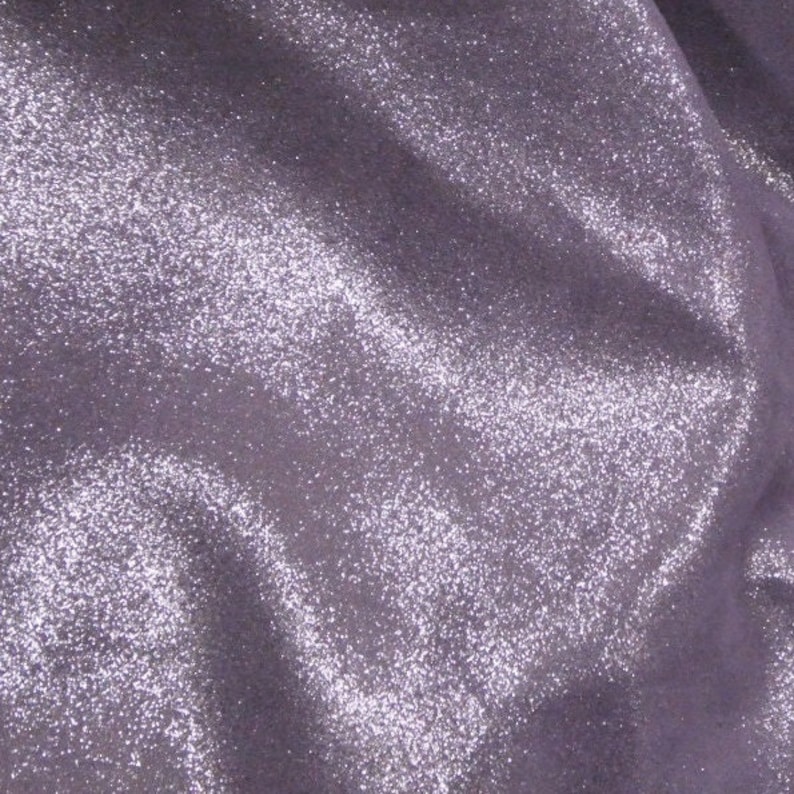 May include: Close-up of a shimmering, lavender fabric. The material is covered in tiny, sparkling glitter, creating a textured, reflective surface. The fabric is draped, with soft folds and shadows.