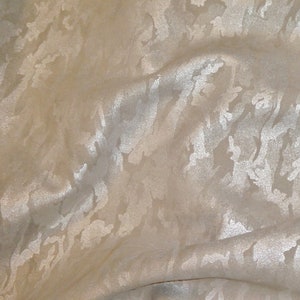 May include: A beige fabric with a white, abstract, camouflage pattern. The fabric has a subtle sheen and a slightly textured surface.