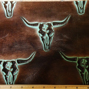May include: Brown leather with a repeating pattern of turquoise-accented bull skull designs. The skulls are embossed into the leather, creating a textured surface. The design is reminiscent of Western or Southwestern decor.