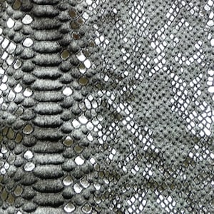 Könnte beinhalten: Nahaufnahme eines strukturierten Stoffes mit Schlangenmuster. Das Design zeigt eine dunkelgraue Basis mit silberfarbenen Schuppen, die einen realistischen Reptilienhaut-Effekt erzeugen. Die Schuppen variieren in Größe und Form und verleihen Tiefe.
