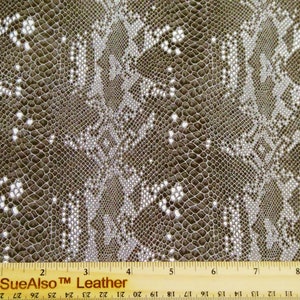 May include: Close-up of a brown and white snakeskin patterned fabric with a ruler measuring 9 inches. The fabric has a textured surface and a repeating pattern.